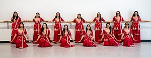 Fourteen women in matching red traditional outfits pose in two rows, standing and kneeling, with hands resting on a ballet barre against a white wall.