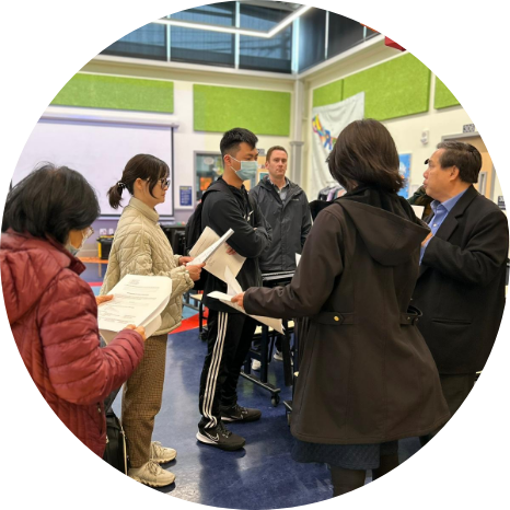 A group of people stand in a classroom, holding papers and having a discussion. Some are wearing jackets and one person is wearing a mask.