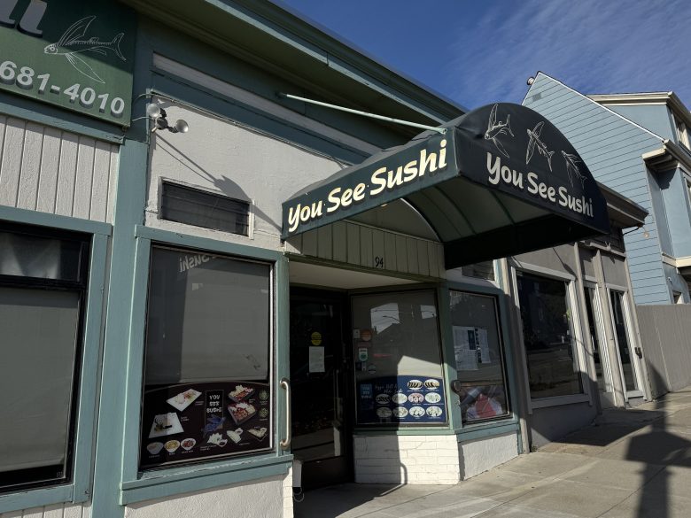 Storefront of "You See Sushi" restaurant with a black awning displaying the name and menu photos on the window, located on a sunny street with neighboring buildings.