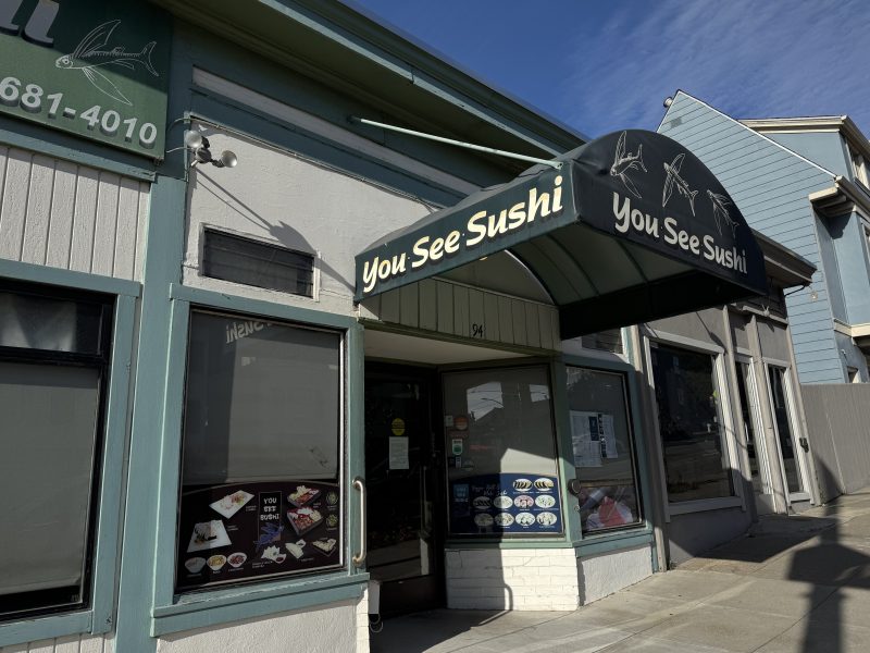 Storefront of "You See Sushi" restaurant with a black awning displaying the name and menu photos on the window, located on a sunny street with neighboring buildings.