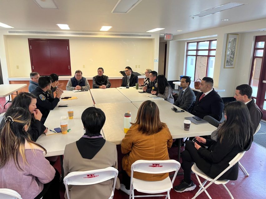 A group of people sit around a large rectangular table in a meeting room, engaged in discussion. Some have drinks in front of them.
