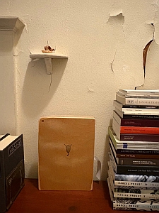 A small shelf with a figurine is mounted on a wall with peeling paint; below, a wooden plank rests next to two stacks of books on a wooden surface.
