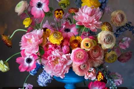 A blue vase holds a colorful arrangement of pink, yellow, and orange flowers with green stems, including roses, ranunculus, and anemones.