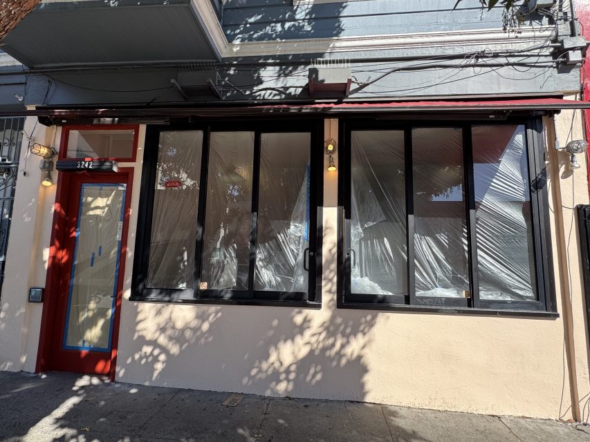 Storefront with large windows covered in plastic sheeting, door taped with paper, and two wall-mounted lights, suggesting renovation or construction in progress.