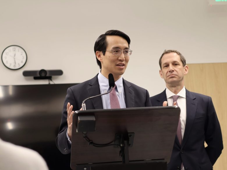 Two men in suits stand at a podium; one speaks into a microphone while the other looks on. A clock, monitor, and webcam are visible in the background.