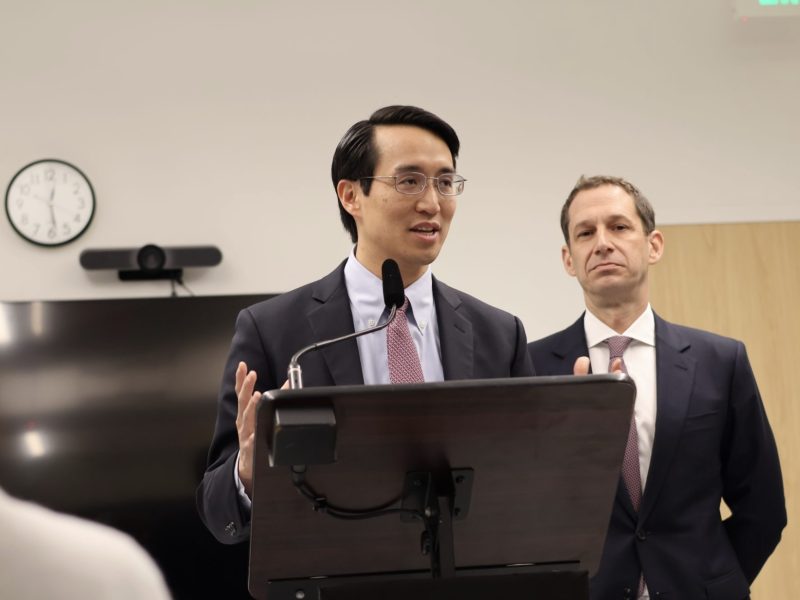 Two men in suits stand at a podium; one speaks into a microphone while the other looks on. A clock, monitor, and webcam are visible in the background.