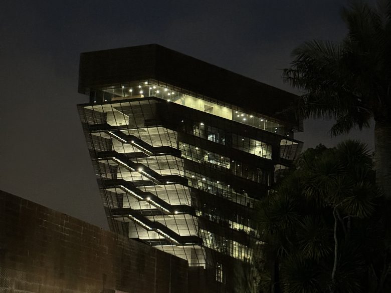 A modern, angular building with illuminated exterior stairs is seen at night, framed by palm trees and a dark sky.