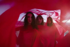 Three women stand together, partially obscured by flowing red fabric, with a soft light in the background.