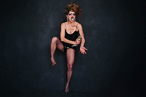 Person wearing a black outfit and high heels poses energetically against a dark background, holding a microphone and singing with an expressive facial expression.