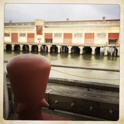 A red bollard in the foreground overlooks a body of water and an old industrial building with multiple closed red doors.