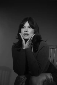A woman with dark hair sits on a wooden chair, resting her face in her tattooed hands, looking slightly upward. The image is in black and white.