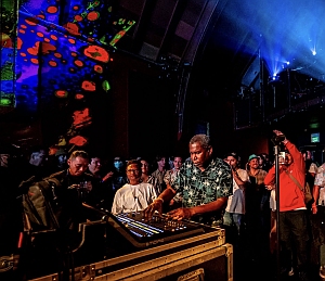 A man operates a soundboard at a concert, surrounded by a crowd; colorful lights and projections illuminate the venue.