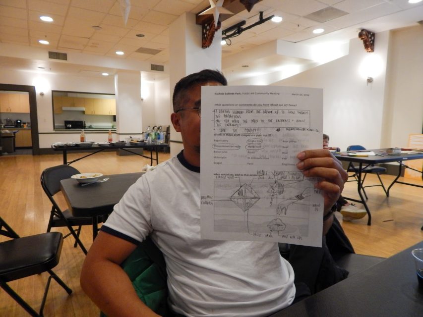 A person sits at a table in a room, holding up a sheet of paper with drawings and writing, partially obscuring their face. Tables, chairs, and snacks are visible in the background.