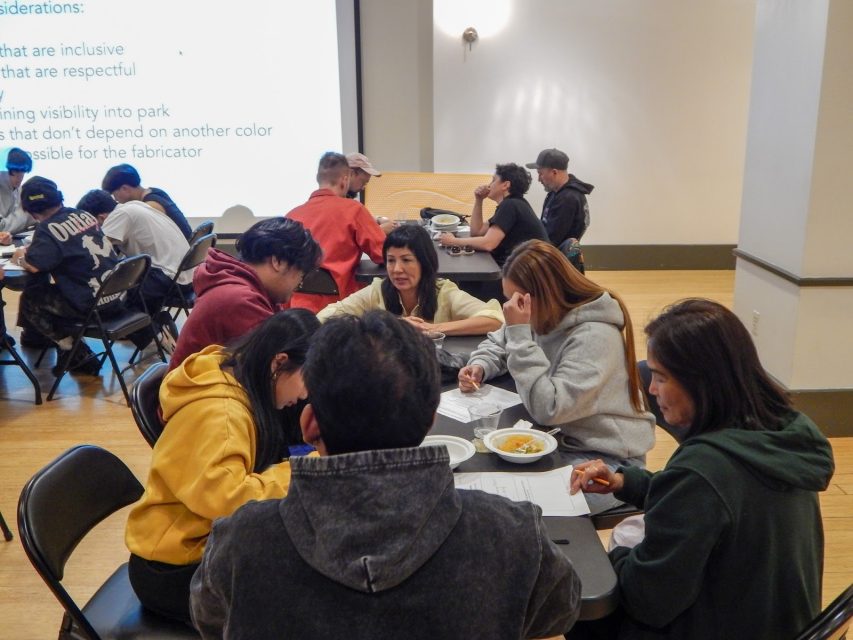 A group of people sit around tables in a classroom, writing on papers and eating, with a presentation projected on a screen in the background.