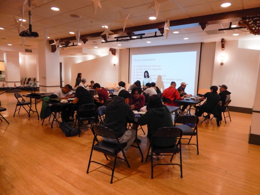 People sit in groups at tables in a spacious room, facing a projector screen displaying a presentation. The atmosphere appears to be that of a workshop or classroom session.