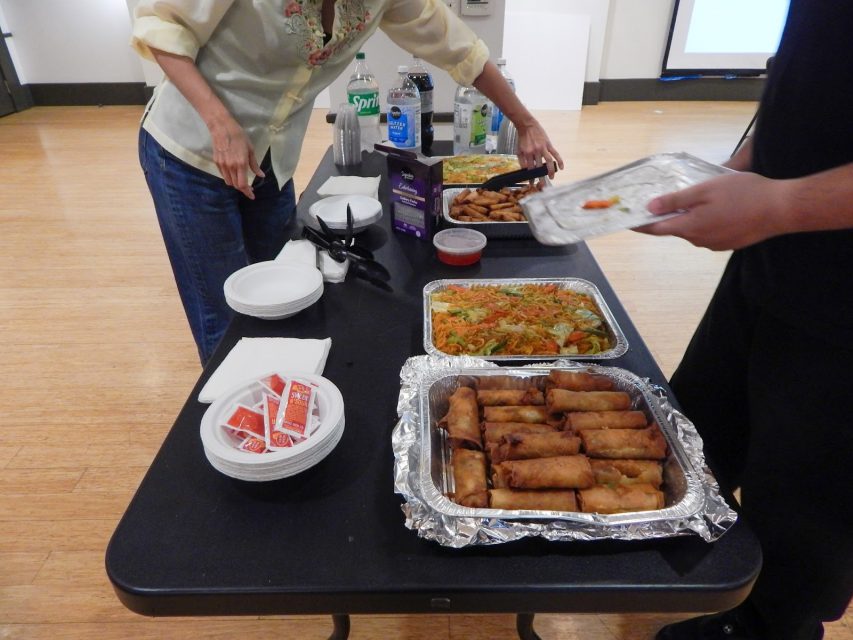 Two people serve food from trays of spring rolls and noodles on a table, with plates, utensils, sauce packets, and bottled drinks also visible.