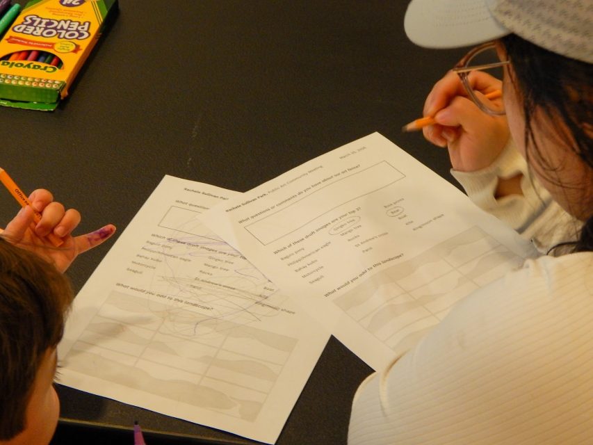 Two people sit at a table coloring on worksheets with crayons; one sheet has scribbles, and a box of Crayola colored pencils is visible in the corner.