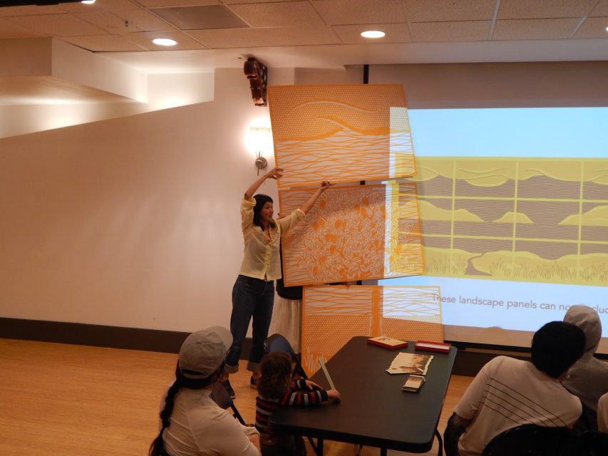 A woman stands in front of a group, holding and discussing large orange landscape art panels, with a presentation projected on the wall behind her.