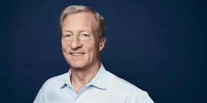A man with short light hair wearing a light blue button-down shirt stands in front of a plain dark blue background, smiling at the camera.