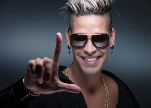A man with spiked blonde hair, sunglasses, earrings, and gold chains smiles while holding up his hand in an "L" shape.