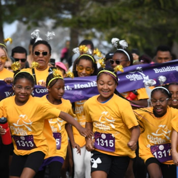 A group of girls in yellow shirts run together at a race event, smiling and holding hands, with adults and a purple banner in the background.