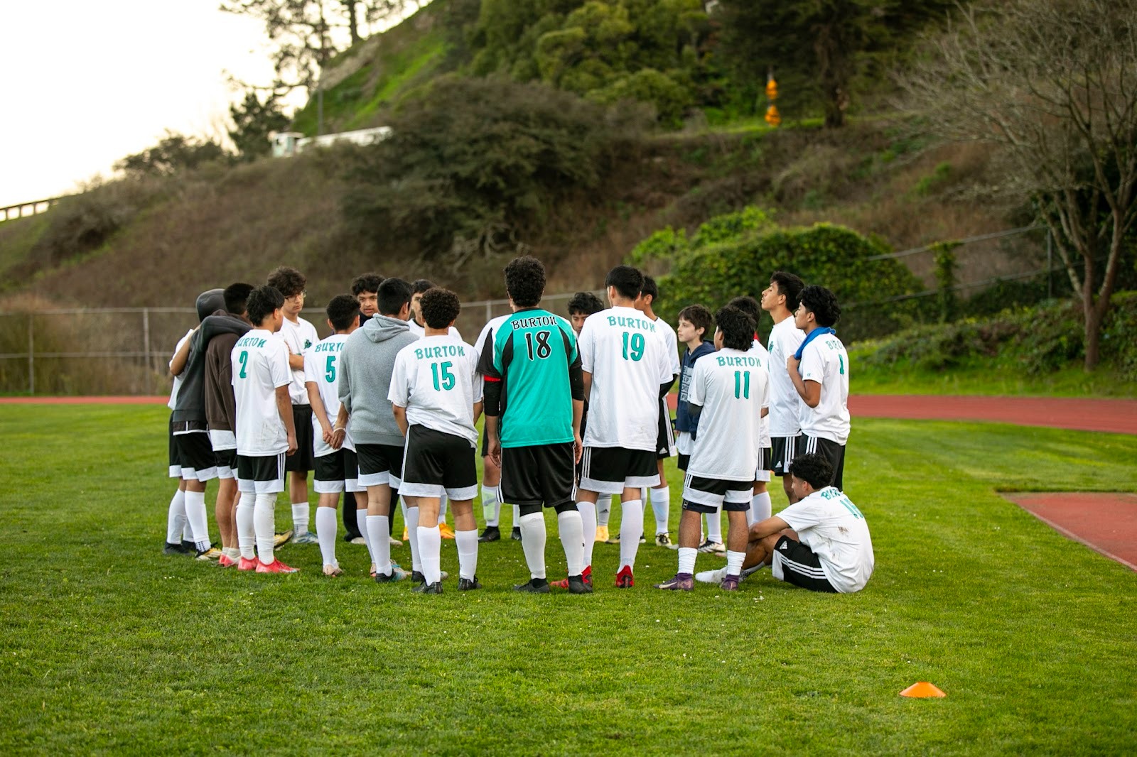 A soccer team in matching uniforms stands in a circle on a grassy field, listening to instructions, with trees and a track visible in the background.
