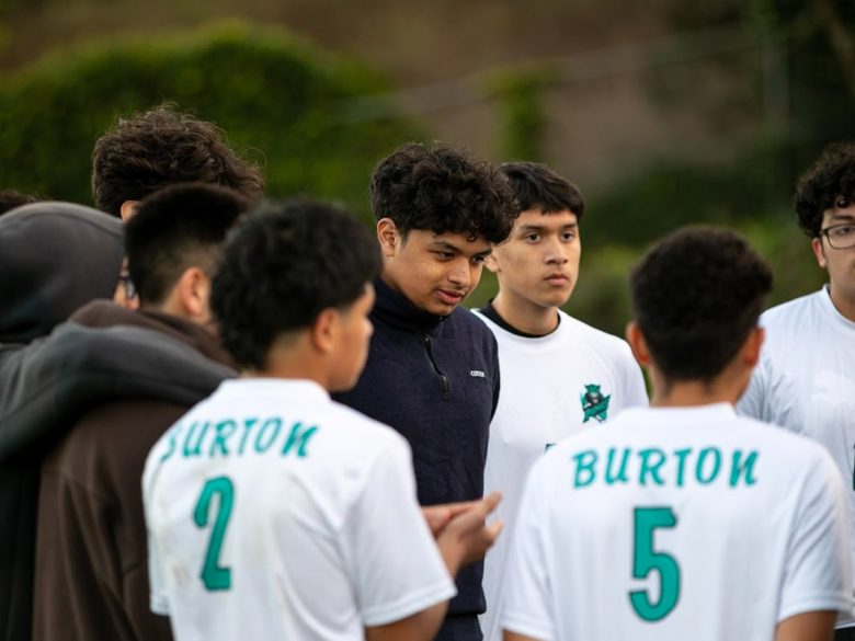A group of teenage boys stand together outdoors; some wear white soccer jerseys labeled "BURTON" with numbers 2 and 5, while others wear casual clothes.