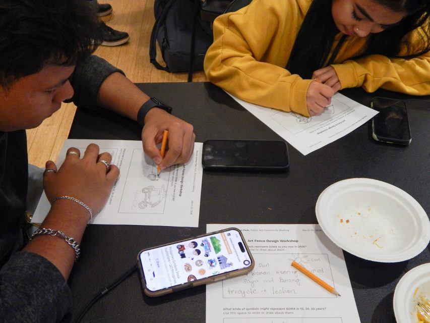 Two people sit at a table working on worksheets with pencils; a phone, empty plate, and papers are on the table.