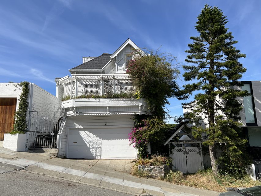 A white two-story house with a balcony, garage, and a tall tree in front, situated on a steeply sloped street under a blue sky.