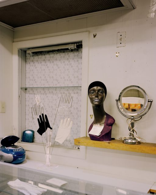 A store display with a mannequin head on a shelf, a mirror, glove stands with gloves, and various items on a glass counter in front of a patterned window.