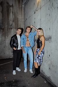 Three people pose against a concrete wall; one man wears a black jacket, another in denim shows his torso, and a woman wears a black top with a patterned skirt and black boots.