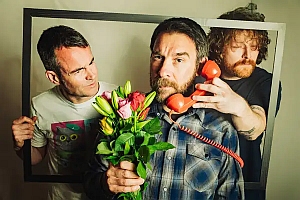 Three men pose with props: one holding a picture frame, another holding flowers, and the third placing a red phone receiver to the central figure’s ear.