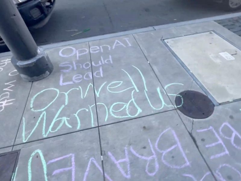 Chalk writing on a sidewalk reads, "OpenAI Should Lead Orwell Warned Us," with additional partially visible words and passing cars nearby.