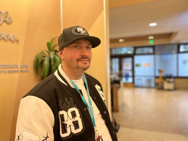 Man wearing a black baseball cap and varsity jacket stands indoors near a plant, with a blurred hallway and dining room in the background.