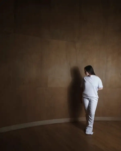 A person dressed in white stands facing a plain brown wall in a dimly lit room with a wooden floor.