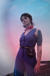 Person standing outdoors at sunset wearing a blue outfit, red bandana, waist bag, and showing a tattoo on their left forearm. The background features a gradient sky and a shoreline.