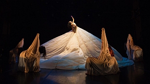 A performer stands atop a large, dramatic, white gown while four figures draped in fabric crouch around the skirt on a dark stage.