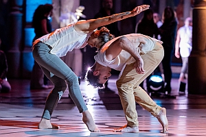 Two barefoot dancers in casual clothing perform a contemporary dance routine, leaning toward each other with outstretched arms on a lit stage.