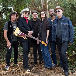 Six adults stand outdoors among trees, posing for a group photo; one person in front holds an electric guitar.