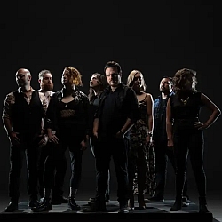 Eight people stand in a dark setting, arranged in a group and facing forward, with neutral to serious expressions.