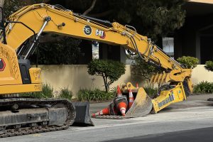 A yellow CAT excavator parked on the street holds several orange traffic cones in its bucket near a sidewalk and greenery.