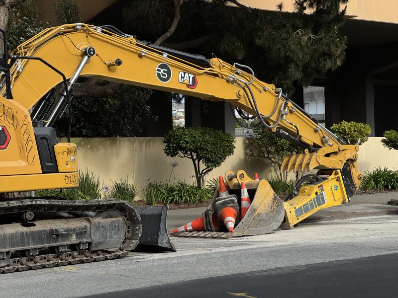 A yellow CAT excavator parked on the street holds several orange traffic cones in its bucket near a sidewalk and greenery.