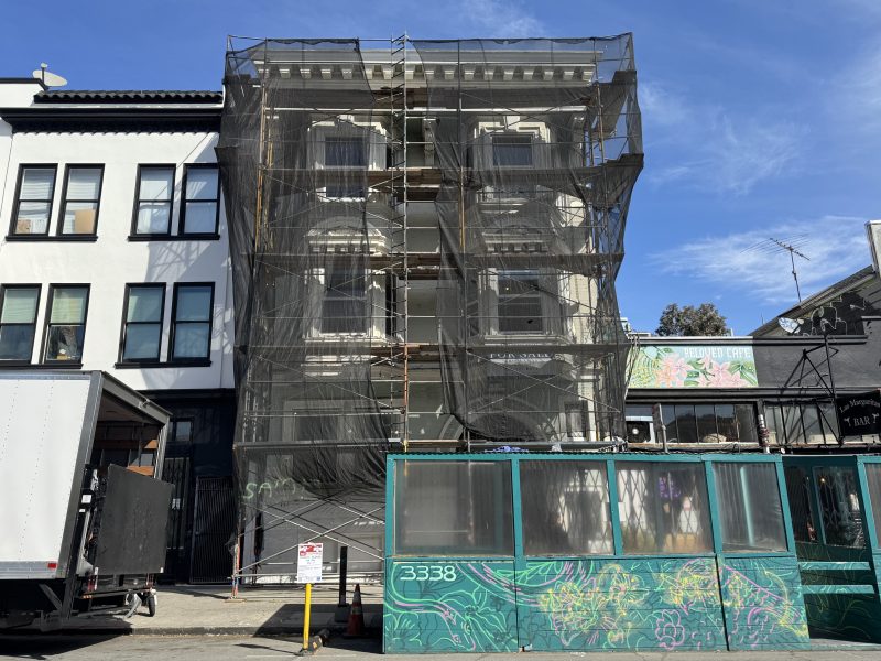 A three-story building under renovation is covered in black construction netting and scaffolding, with a green temporary structure in front and a clear blue sky above.