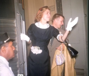 A woman in a black dress and white gloves waves while standing near a train door, accompanied by a man holding a coat and handbag. A uniformed person sits nearby.