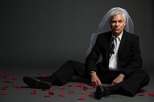 A person in a black suit and white veil sits on the floor with scattered red rose petals, against a dark background.