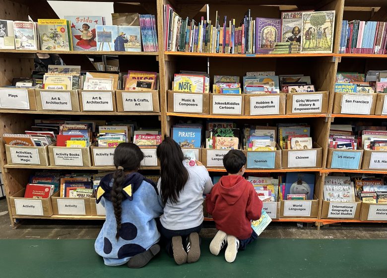 Three children kneel on the floor, browsing books on low shelves in a library section labeled with various genres and subjects.