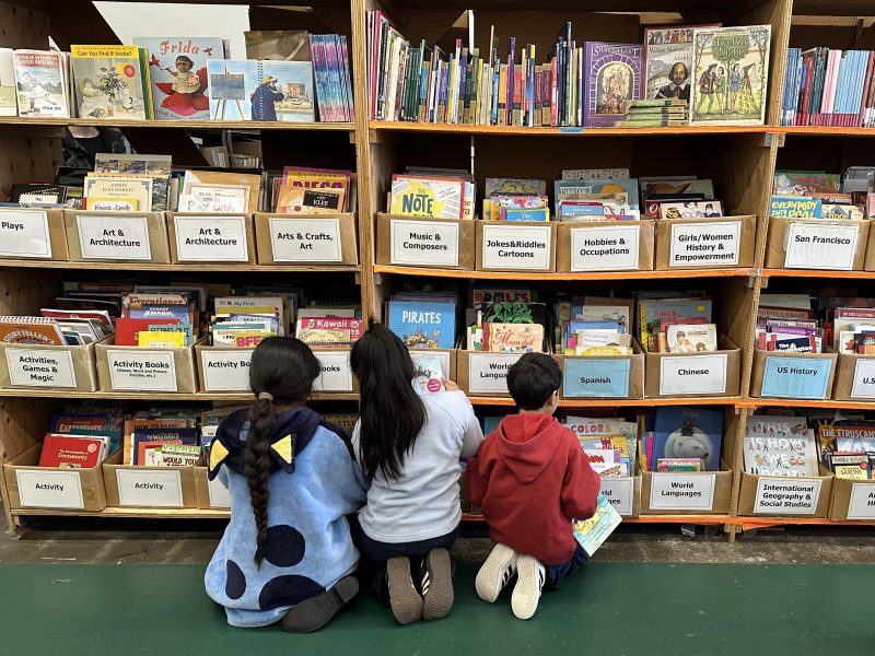 Three children kneel on the floor, browsing books on low shelves in a library section labeled with various genres and subjects.