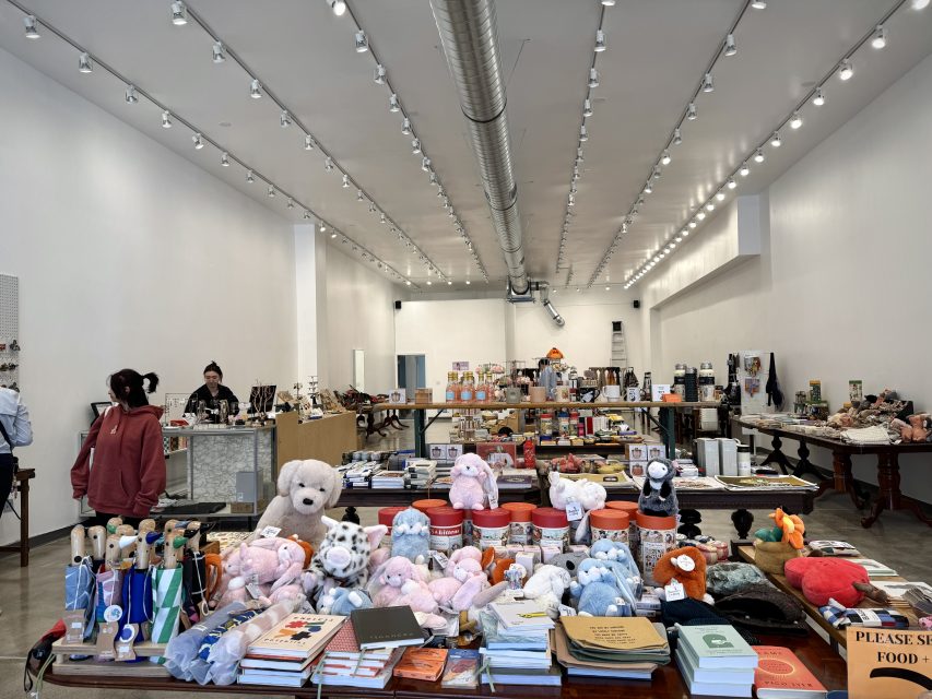 A well-lit gift shop interior with plush toys, books, and various items displayed on tables; a few people are browsing near the checkout counter.