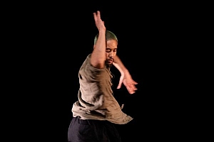 A person with short hair and a loose shirt dances against a black background, with one arm raised and eyes closed.