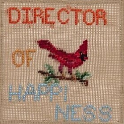 A cross-stitched design showing a red cardinal on a branch, with the words "Director of Happiness" stitched in colorful letters.